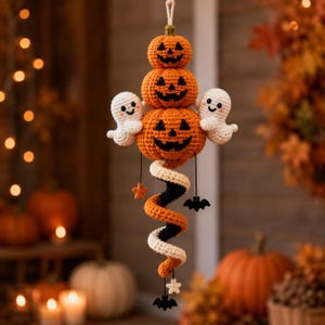 May include: A hanging crochet decoration featuring three stacked orange jack-o'-lanterns with black stitched faces, flanked by two small white ghosts. A spiral tail in orange, black, and white hangs below, accented with small black bats and white stars.
