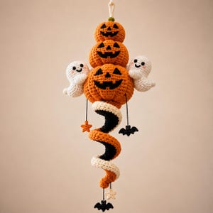 May include: A hanging crochet decoration featuring three stacked orange jack-o'-lanterns with black faces, flanked by two small white ghosts. A spiral tail in orange, black, and white extends downward, accented with small black bats and stars.