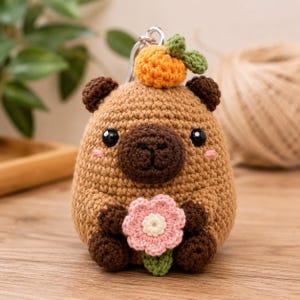 Puede incluir: Un llavero de capibara de ganchillo con cuerpo color canela, orejas y hocico marrón oscuro, y ojos de plástico negro. El capibara tiene una pequeña fruta naranja con una hoja verde en la cabeza y sostiene una flor rosa con centro blanco.