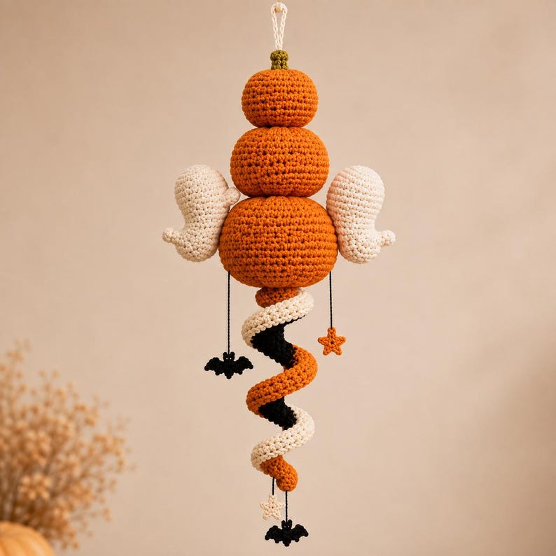 May include: A hanging crochet decoration featuring three stacked orange pumpkins topped with a small green stem. Two white crocheted ghosts extend from the sides. Below, a spiral tail in orange, black, and white hangs with small dangling black bats and orange and white stars.