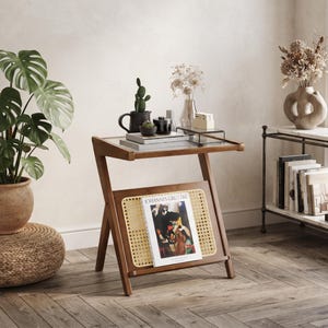 May include: A Z-shaped wooden side table with a glass top and a lower rack featuring a woven cane panel. A book titled Johannes Grützke is resting against the cane section. The table has a warm brown finish and a clean, angular frame design.