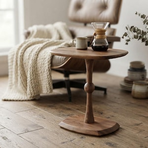 Puede incluir: Una mesa auxiliar de madera con una superficie lisa y redondeada y una base de pedestal central con un detalle esférico. La mesa está hecha de madera de tono medio con veta visible, apoyada sobre una base ovalada plana en el suelo.