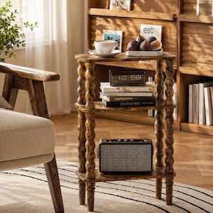 May include: A three-tiered wooden side table with turned, spindle-style legs. The top shelf holds a white ceramic cup on a saucer and a small tray of figs. The middle shelf contains a digital clock and stacked books. A speaker sits on the bottom shelf.