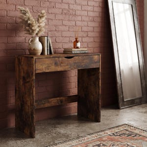 May include: A dark brown wooden console table with a single drawer featuring a cutout handle. The table surface holds a ceramic vase with dried pampas grass, two stacked books, and a small amber glass bottle with reed diffuser sticks.