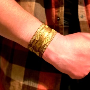 May include: A wide gold-toned metal cuff bracelet featuring a hammered texture and intricate engraved geometric patterns, including horizontal bands of repeating chevron and linear motifs that wrap around the entire surface of the wrist.