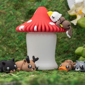 May include: A white mushroom-shaped container with a red cap featuring white spots. A small bald eagle figure rests on the cap. Four small animal figures, including a black cat, a deer, a platypus, and a raccoon, are positioned at the base.
