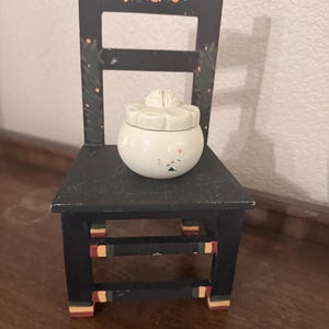 May include: A small, black wooden chair with hand-painted yellow floral accents on the backrest and colorful stripes on the legs. Resting on the seat is a round, off-white ceramic jar with a lid shaped like a flower, featuring a small paint chip.