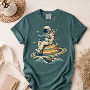 Puede incluir: Una camiseta de cuello redondo en color verde azulado oscuro con un gráfico de un astronauta sentado sobre un planeta con anillos, leyendo un libro azul con una taza de café humeante al lado. El diseño incluye estrellas amarillas y nubes.