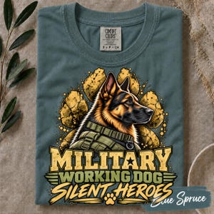 May include: A slate blue t-shirt featuring a graphic of a German Shepherd wearing a tactical vest. The dog is centered over a yellow paw print design. Below the dog, the text reads "MILITARY WORKING DOG SILENT HEROES" in bold lettering.