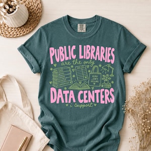 May include: A dark teal short-sleeve t-shirt featuring the pink text "PUBLIC LIBRARIES" and "DATA CENTERS" with green line art illustrations of a stack of books, a library card, a tote bag, a lamp, a plant, and an open book with the text "i support".