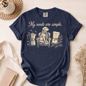 Puede incluir: Una camiseta azul marino de manga corta con el texto "My needs are simple." El gráfico incluye una cafetera italiana, granos de café, un perro con bufanda sentado en un sillón, un libro titulado "The Literary Journal", una pluma y gafas.