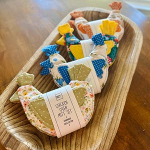 May include: A set of three fabric chicken-shaped oven mitts with floral and polka dot patterns in green, blue, and orange. Each mitt features a white paper band with the text CHICKEN OVEN MITT SET HANDMADE 100% COTTON, arranged in a wooden bowl.