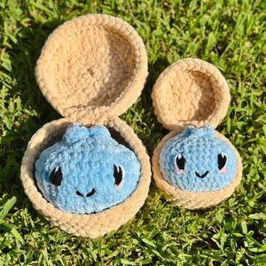 May include: Two handmade crochet plushies, each featuring a light blue dumpling character with black eyes and a small smile, nestled inside a tan, circular, open-topped container. The plushies are made of soft, textured yarn with a fuzzy appearance.