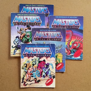 May include: A collection of vintage "Masters of the Universe" mini-comics. The covers feature vibrant illustrations of characters and scenes, with titles like "King of Castle Grayskull" and "He-Man and the Power Sword". The comics are arranged on a light brown surface.