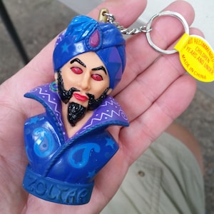 May include: A blue and purple plastic keychain featuring a caricature of a genie with a beard and a turban. The keychain has the word "ZOLTAR" written on the bottom. The keychain has a warning label that reads "Not recommended for children 3 years and under. Made in China."
