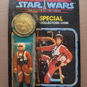 Op de afbeelding: Vintage Star Wars actiefiguur van Luke Skywalker in X-Wing pilootuitrusting, verpakt op een kaart met een goudkleurige verzamelaarsmunt. De kaart toont de tekst "STAR WARS THE POWER OF THE FORCE" en "SPECIAL COLLECTORS COIN". Geschikt voor kinderen vanaf 4 jaar.