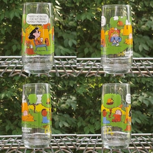 May include: Four clear glass tumblers with colorful illustrations of Peanuts characters camping. Each glass features a different scene with text, including "There's no excuse for not being properly prepared."