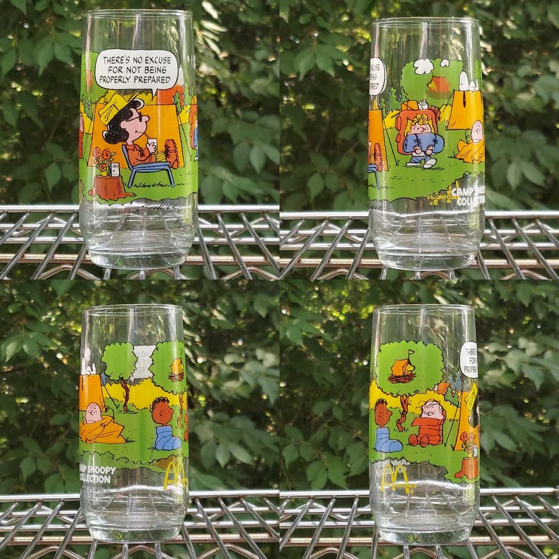 May include: Four clear glass tumblers with colorful illustrations of Peanuts characters camping. Each glass features a different scene with text, including "There's no excuse for not being properly prepared."