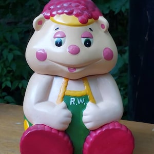 May include: A ceramic cookie jar shaped like a cartoon squirrel wearing a green and yellow outfit. The jar has a red and yellow hat and the words "R.W." on the front.