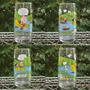 May include: Four clear glass tumblers with colorful cartoon illustrations of Snoopy and the Peanuts gang. Each glass features a different scene with characters in a river setting. One glass has the text "RATS! WHY IS HAVING FUN ALWAYS SO MUCH WORK?" and another has the text "CAMP SNOOPY COLLECTION".