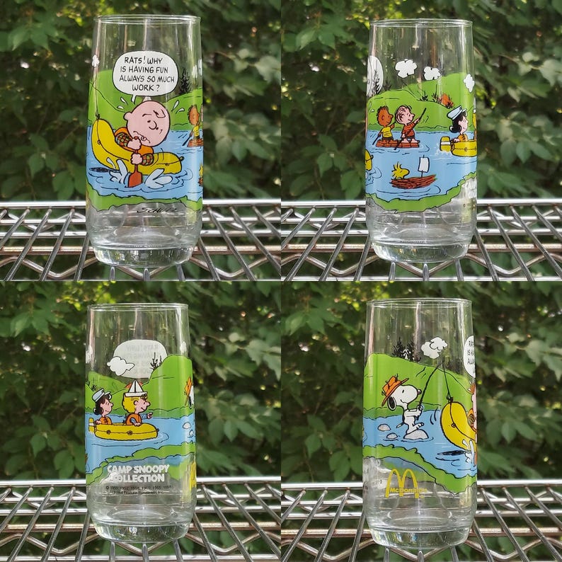 May include: Four clear glass tumblers with colorful cartoon illustrations of Snoopy and the Peanuts gang. Each glass features a different scene with characters in a river setting. One glass has the text "RATS! WHY IS HAVING FUN ALWAYS SO MUCH WORK?" and another has the text "CAMP SNOOPY COLLECTION".