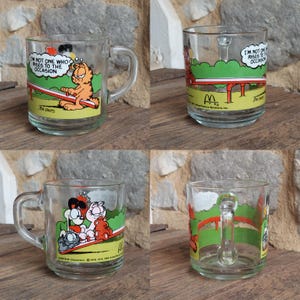 May include: Set of four Garfield themed glass mugs. Each mug features a different Garfield cartoon illustration. The mugs are clear glass with a white rim and handle. The text on the mugs reads "I'm not one who rises to the occasion".