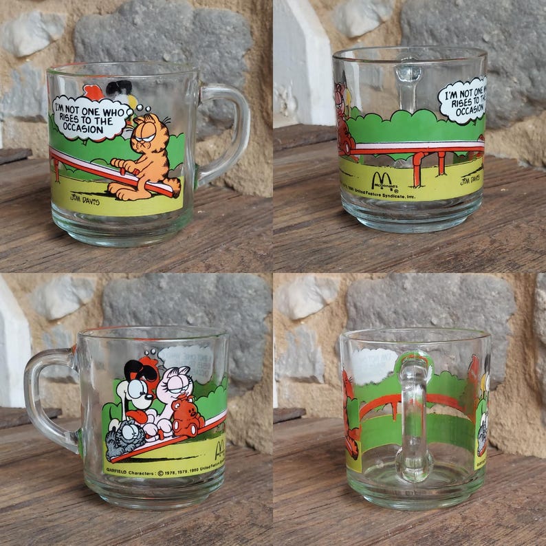May include: Set of four Garfield themed glass mugs. Each mug features a different Garfield cartoon illustration. The mugs are clear glass with a white rim and handle. The text on the mugs reads "I'm not one who rises to the occasion".