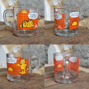 May include: Set of four glass mugs featuring Garfield the cat. Each mug has a different image of Garfield with a sunrise or sunset and a caption. The captions are "I've never seen a sunrise... I'm waiting for the movie", "Such a beautiful sunset... and me without a horse", "I've never seen a sunrise... I'm waiting for the movie", and "Such a beautiful sunset... and me without a horse".