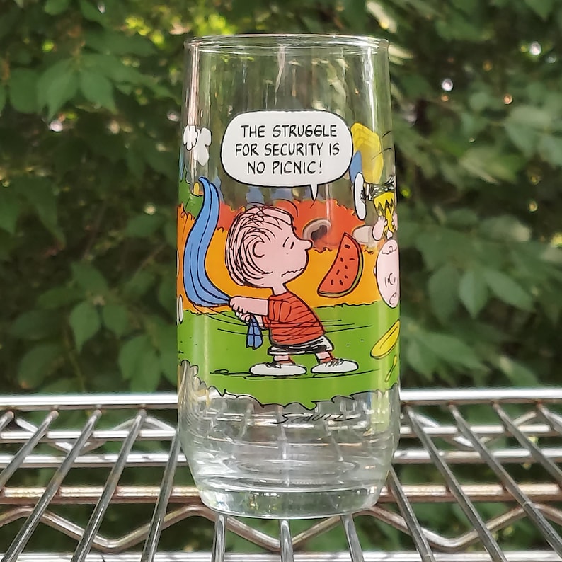May include: A clear glass featuring a cartoon illustration of Charlie Brown from the Peanuts comic strip. The glass has the phrase "THE STRUGGLE FOR SECURITY IS NO PICNIC!" and depicts Charlie Brown with a blue blanket and a slice of watermelon.