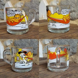 May include: Set of four Garfield themed glass mugs. The mugs feature different illustrations of Garfield, including him relaxing in a hammock, wearing a cape, and with a glass of juice. The mugs have a white background with orange, yellow, and red accents. The text "It's not a pretty life but somebody has to live it" is printed on one of the mugs.