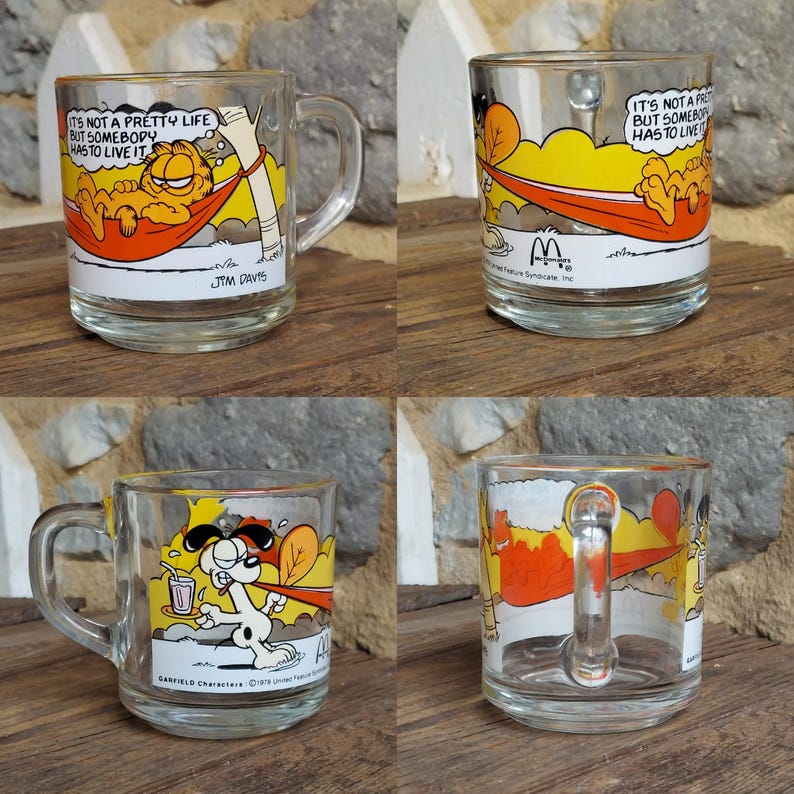 May include: Set of four Garfield themed glass mugs. The mugs feature different illustrations of Garfield, including him relaxing in a hammock, wearing a cape, and with a glass of juice. The mugs have a white background with orange, yellow, and red accents. The text "It's not a pretty life but somebody has to live it" is printed on one of the mugs.