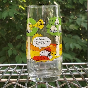 May include: A clear glass featuring a Snoopy cartoon design. The glass has a yellow and green forest scene with Snoopy sleeping on a red pillow. Text includes "BLAAT!" and "MORNING PEOPLE ARE HARD TO LOVE."