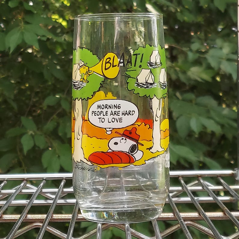 May include: A clear glass featuring a Snoopy cartoon design. The glass has a yellow and green forest scene with Snoopy sleeping on a red pillow. Text includes "BLAAT!" and "MORNING PEOPLE ARE HARD TO LOVE."