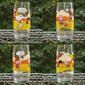 May include: Four clear glass tumblers with colorful cartoon illustrations of Snoopy and other Peanuts characters. Each glass features a different scene with yellow, orange, and white backgrounds. One glass has the text "CIVILIZATION IS OVERRATED!"