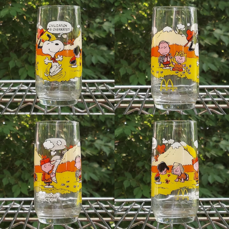 May include: Four clear glass tumblers with colorful cartoon illustrations of Snoopy and other Peanuts characters. Each glass features a different scene with yellow, orange, and white backgrounds. One glass has the text "CIVILIZATION IS OVERRATED!"