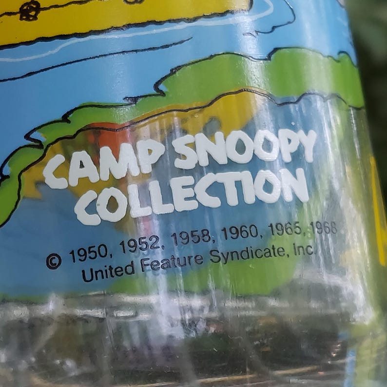 May include: Close-up of a clear glass object with the text "CAMP SNOOPY COLLECTION" in white. The image also includes the years 1950, 1952, 1958, 1960, 1965, and 1968, along with "United Feature Syndicate, Inc."