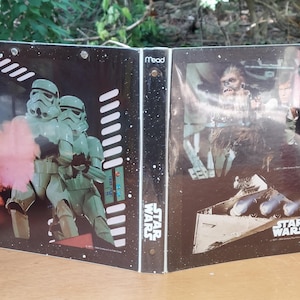 May include: A Star Wars themed binder featuring images of Stormtroopers, Chewbacca, and other characters. The binder is black with white accents and the Star Wars logo. The binder is open and sitting on a wooden surface.