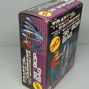 May include: A box for a Transformers 36-inch Bop Bag. The box is purple and black with a 3D image of the Transformers logo. The box has a picture of a child punching the bag. The text on the box reads "Transformers 36" Bop Bag" and "More Than Meets The Eye!"