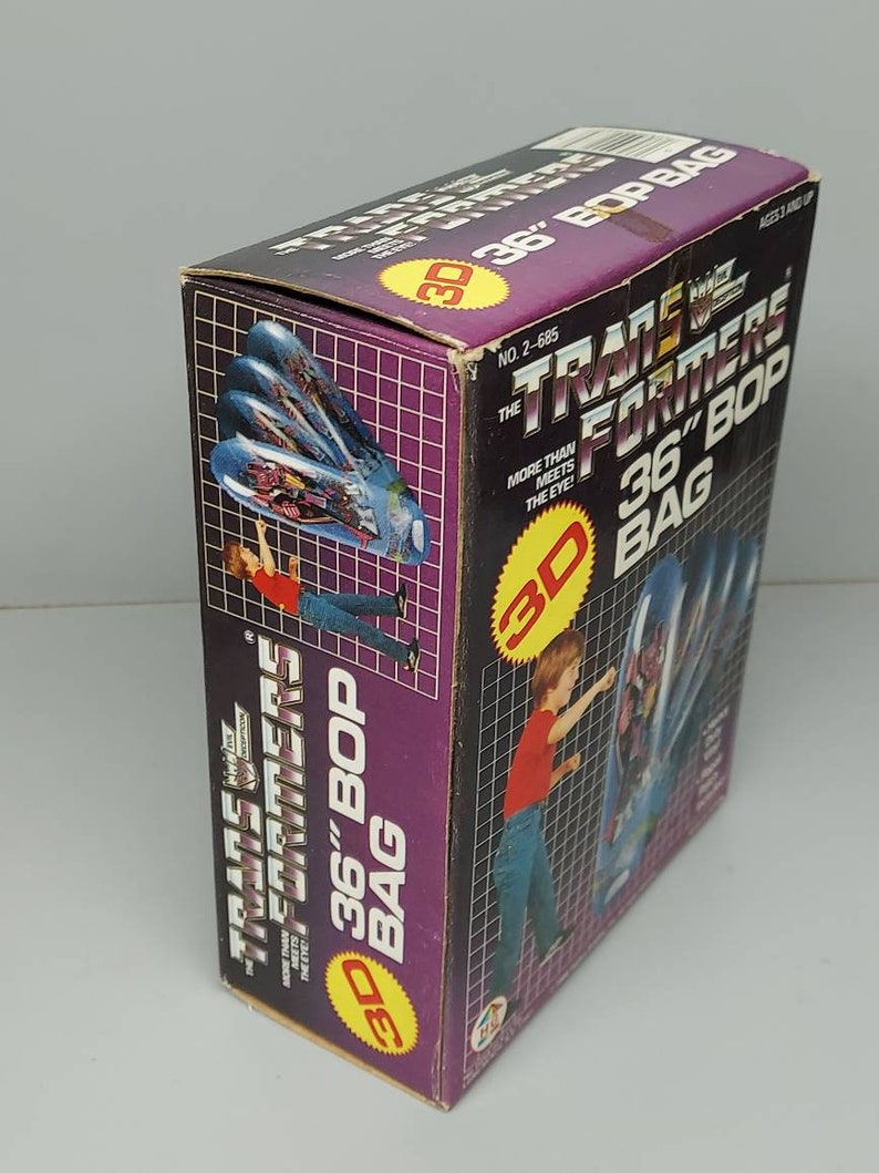 May include: A box for a Transformers 36-inch Bop Bag. The box is purple and black with a 3D image of the Transformers logo. The box has a picture of a child punching the bag. The text on the box reads "Transformers 36" Bop Bag" and "More Than Meets The Eye!"