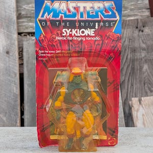 May include: A vintage Sy-Klone action figure from Masters of the Universe, still in its original packaging. The card features the text "NEW!" and "SY-KLONE Heroic fist-flinging tornado". The figure is green, yellow, and orange.
