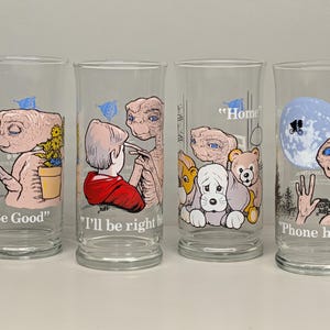 May include: Set of four clear glass tumblers featuring illustrations of E.T. the Extra-Terrestrial. Each glass has a different scene with text: "Be Good", "I'll be right", "Home", and "Phone home". The glasses have a vintage, cartoon style.
