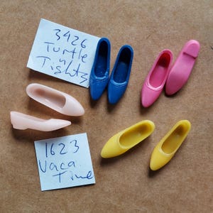 May include: A collection of six pairs of doll shoes in pink, blue, and yellow. The shoes are labeled with the numbers 3426 and 1623. The text "Turtle Tights" and "Vaca Time" are also visible.