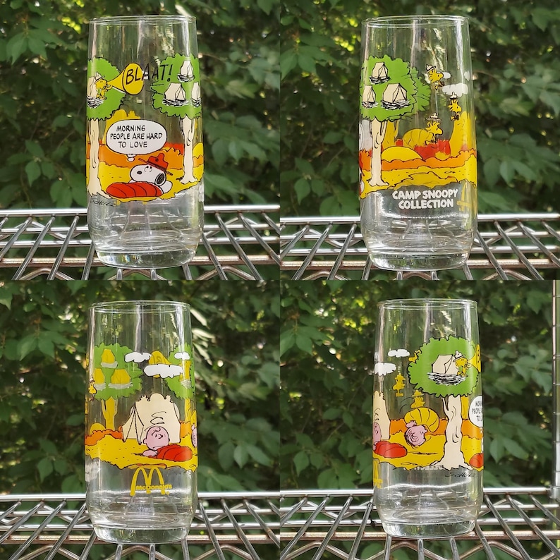 May include: Set of four clear glass tumblers featuring Snoopy and Woodstock camping scenes. Each glass has a different design with text like "BLAAT!" and "Morning people are hard to love." One glass reads "Camp Snoopy Collection."