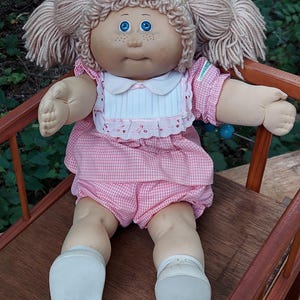 Peut inclure: Une poupée Cabbage Patch Kids vintage avec des couettes blondes en fil, des yeux bleus et des taches de rousseur. La poupée porte une robe vichy rose avec un col blanc et un bloomer assorti. Elle est assise sur une chaise en bois.