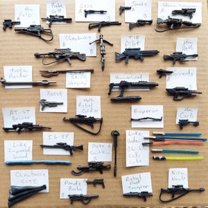 May include: A collection of 24 Star Wars action figures, each featuring a different character and weapon. The figures are arranged in rows on a brown surface. The characters include Han Solo, Boba Fett, Leia Organa, Jawas, Stormtrooper, Chewbacca, TIE Pilot, Han Hoth, Tusken Raider, Lando Skiff, Greedo, Hmmerhead, AT-ST Driver, Bossk, Hoth Rebel Soldier, Emperor, Har Carb, Luke, IG-88, Ponda Baba, Pote Suitkin, Luke Vader, Rebel Fleet Trooper, Nien Numb.