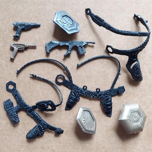 May include: A set of black plastic accessories for a costume or action figure. The set includes two belts, two shields, a gun, and a smaller gun. The accessories are all detailed with intricate designs.
