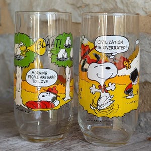 Può includere: Due bicchieri di vetro trasparente con illustrazioni di Snoopy dei fumetti Peanuts. Un bicchiere presenta Snoopy con il testo "BLAAT!" e "MORNING PEOPLE ARE HARD TO LOVE". L'altro bicchiere ha Snoopy con il testo "CIVILIZATION IS OVERRATED!"