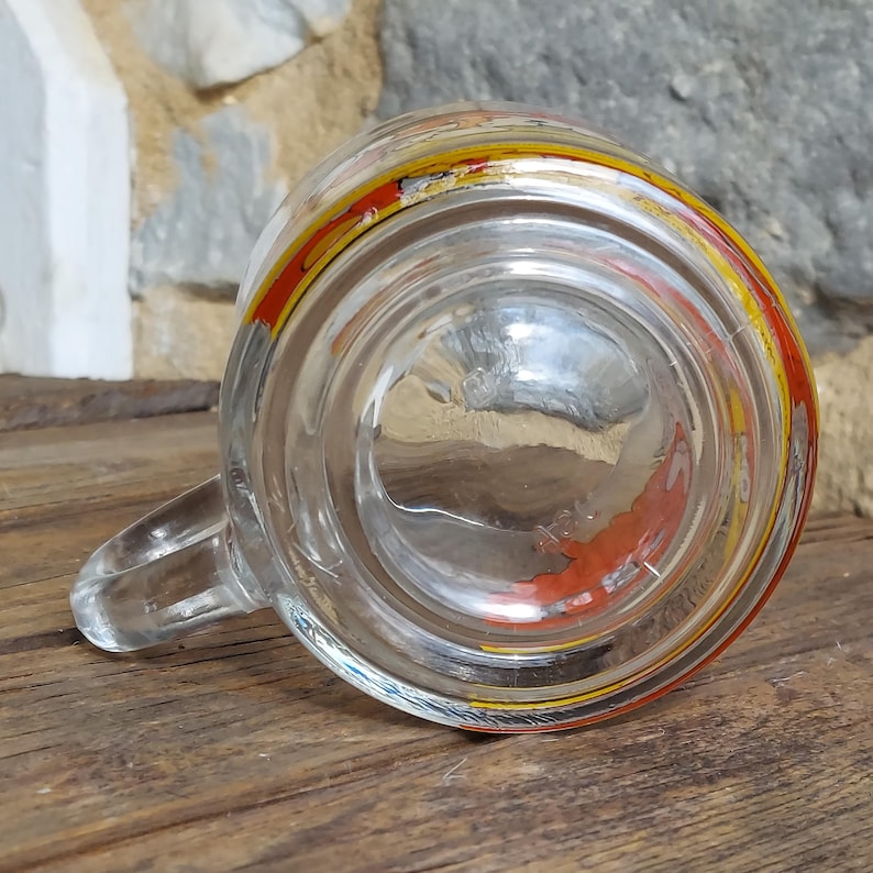 May include: A clear glass mug with a yellow and red rim and a red design on the bottom. The mug has a handle.