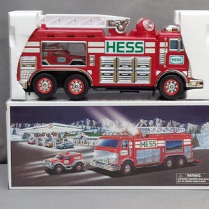 May include: A red Hess toy fire truck with a ladder, lights, and the Hess logo on the side. The truck is in its original box, which features a winter scene with a Hess gas station and a smaller toy truck.