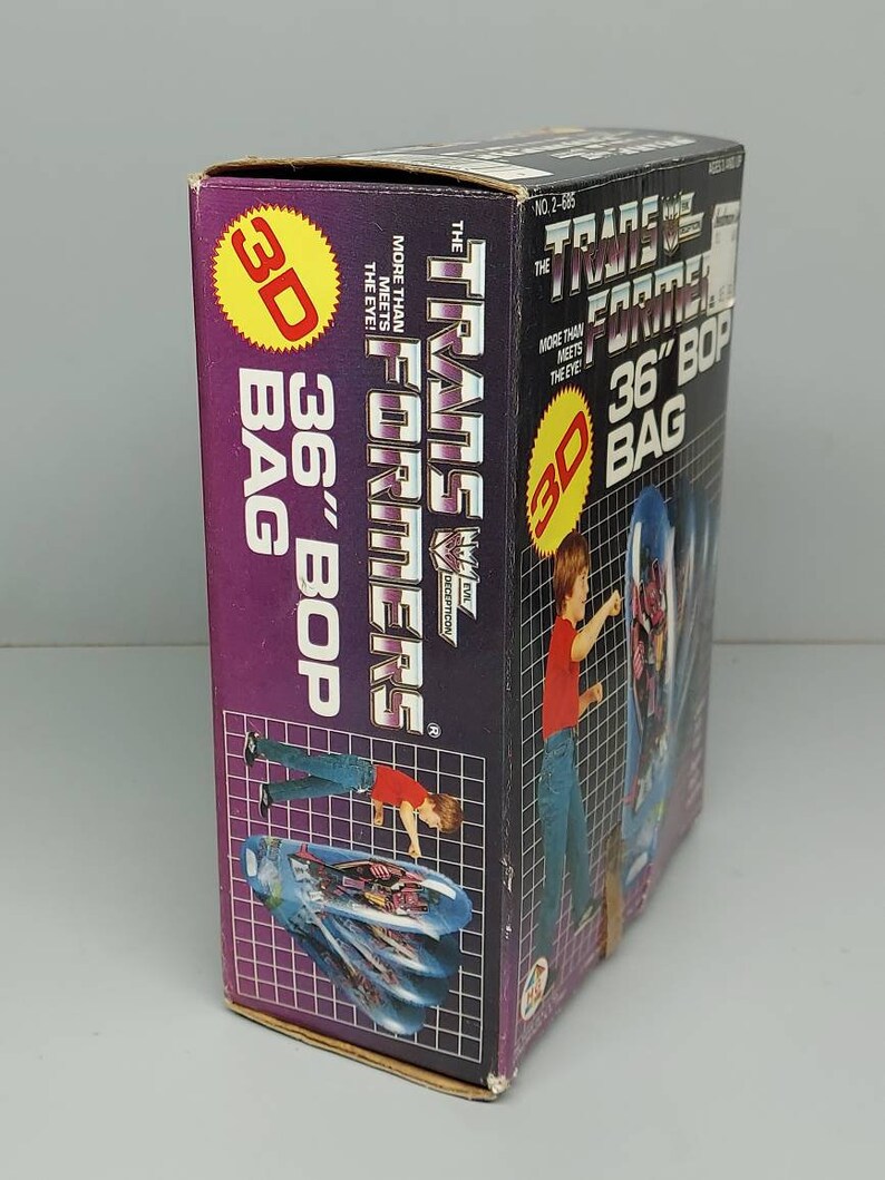 May include: A cardboard box for a Transformers 36-inch Bop Bag. The box is purple and features a 3D image of the Transformers logo and a robot. The box also has the text "The Transformers" and "More Than Meets The Eye!" printed on it.